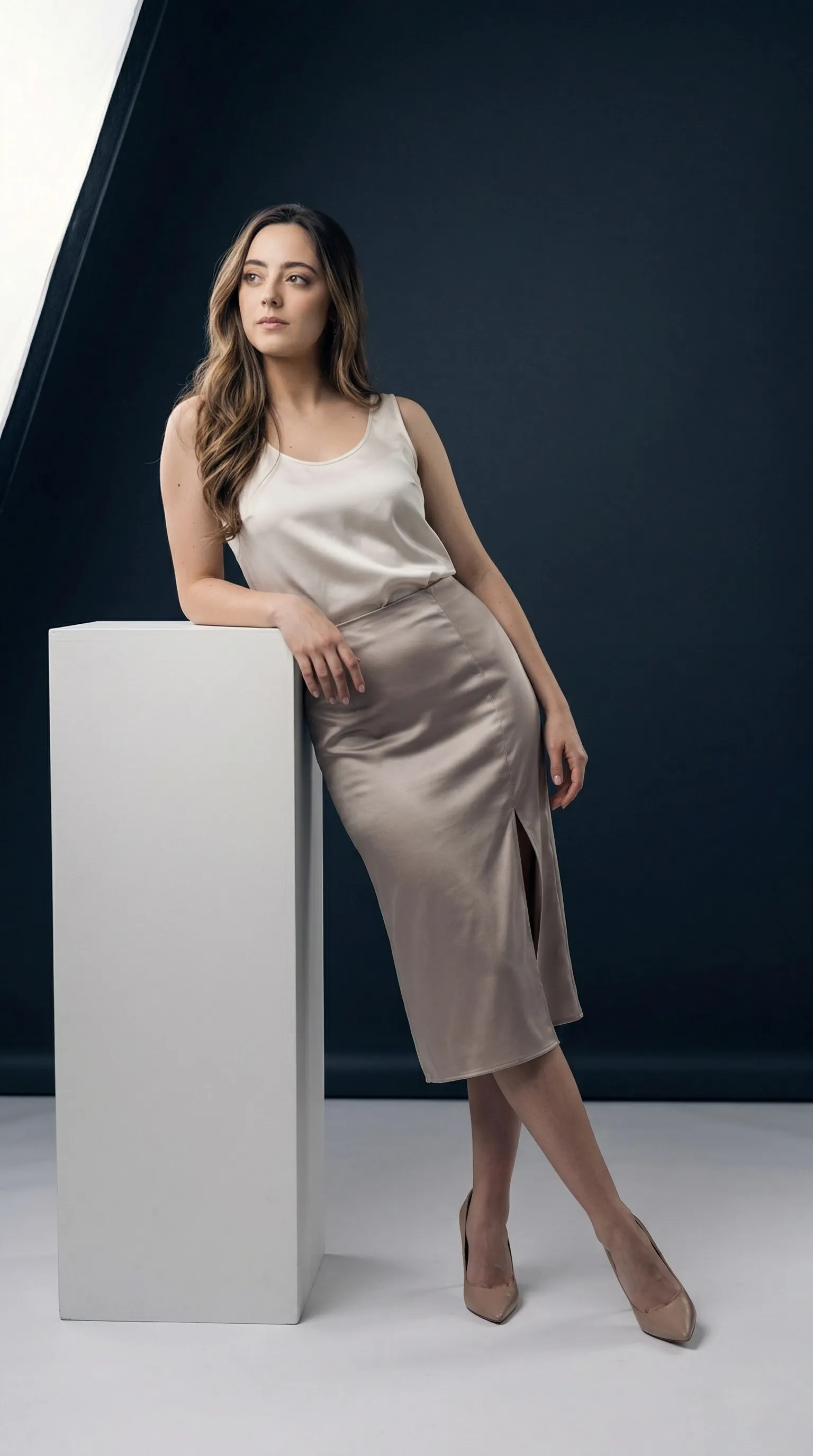 Use the face of the woman in the image provided as inspiration in order to render Ultra–high-resolution cinematic studio portrait designed with the visual tone of a prestige Hollywood character poster. Vertical composition. Clean final render with no text, logos, branding, UI elements, arrows, or interface indicators anywhere in the frame. Minimalist studio environment with a smooth, deep charcoal-to-navy gradient backdrop that transitions subtly from darker tones on camera-right to slightly lighter tones toward camera-left. The background should appear seamless and continuous with no visible edges or corners, creating a controlled editorial studio atmosphere. Floor surface is matte white with soft light reflection, blending naturally into the studio environment. A tall rectangular white pedestal block stands in the foreground slightly left of center. Its surface is matte and clean with crisp edges, functioning as the only prop in the scene. The subject leans casually against this pedestal with the forearm resting on the top edge while the body angles slightly toward camera-right.Pose: relaxed yet confident editorial stance. One hip gently rests against the pedestal while the upper torso leans lightly into the supporting arm. The opposite arm rests softly near the waistline with the hand relaxed. Legs are elegantly crossed at the ankles with one leg extended slightly forward, creating a long diagonal body line. The head turns gently toward camera-left with the gaze directed slightly upward and outward, not directly at the camera, conveying a thoughtful and composed cinematic attitude.
Wardrobe styling: elegant sleeveless top paired with a refined satin-like skirt featuring a side slit, combined with pointed high-heel footwear. Fabric textures should subtly catch the studio light without producing harsh reflections.Lighting (replicate carefully):
Primary soft key light positioned camera-left using a large diffused softbox at slightly above head height. This light produces smooth illumination across the front plane of the body and pedestal with gentle shadow falloff toward camera-right. A subtle fill light from camera-right prevents deep shadow while maintaining dimensional contrast. Very soft rim lighting from behind lightly separates the subject from the dark background, creating a cinematic outline.