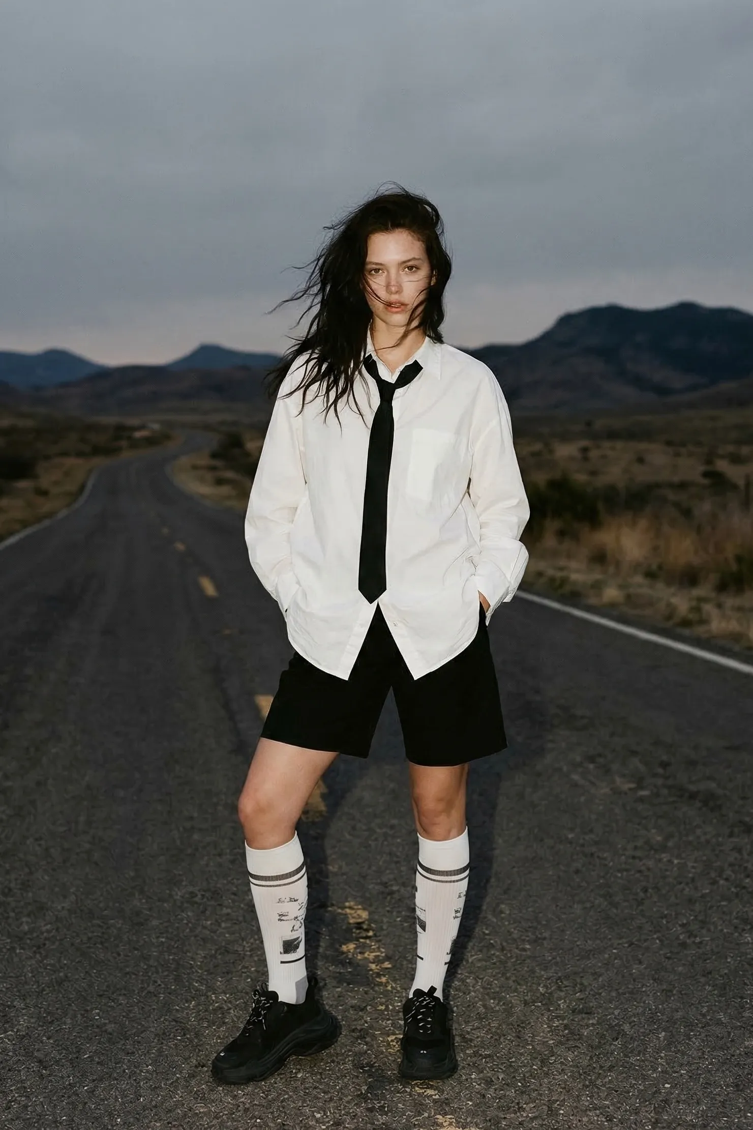 A full-body shot captures a young woman with light skin and long, dark, tousled hair, standing confidently on a deserted road at dusk. She wears an oversized white shirt and a loosened black tie, paired with black shorts and knee-high white socks with graphic print. Her expression is fierce, with intense eye contact and hair blowing across her face. The setting is minimalist, with a winding road disappearing into a hilly, silhouetted landscape under an overcast sky, leading the eye from front to back. The lighting is flat and natural, casting no strong shadows. The color palette is mainly muted with deep blacks and neutral tones. --ar 2:3 --raw --stylize 150